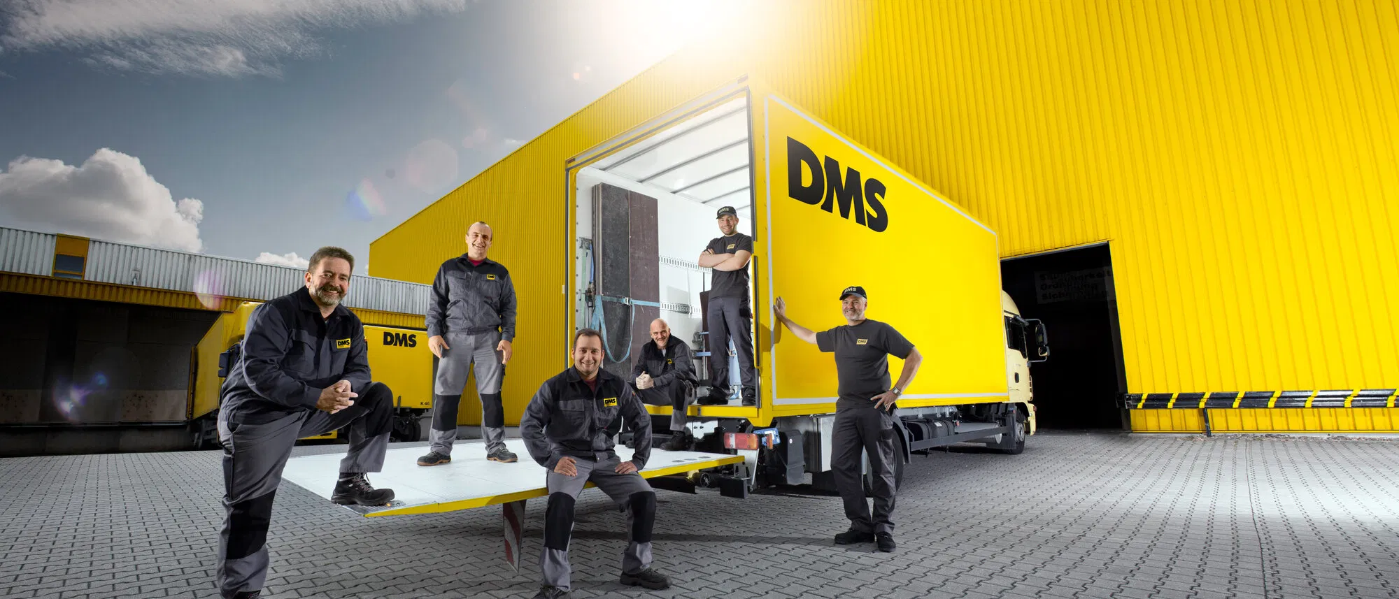 Sechs Mitarbeiter von Bartsch & Weickert am geöffneten gelben Möbelwagen vor einer Lagerhalle.